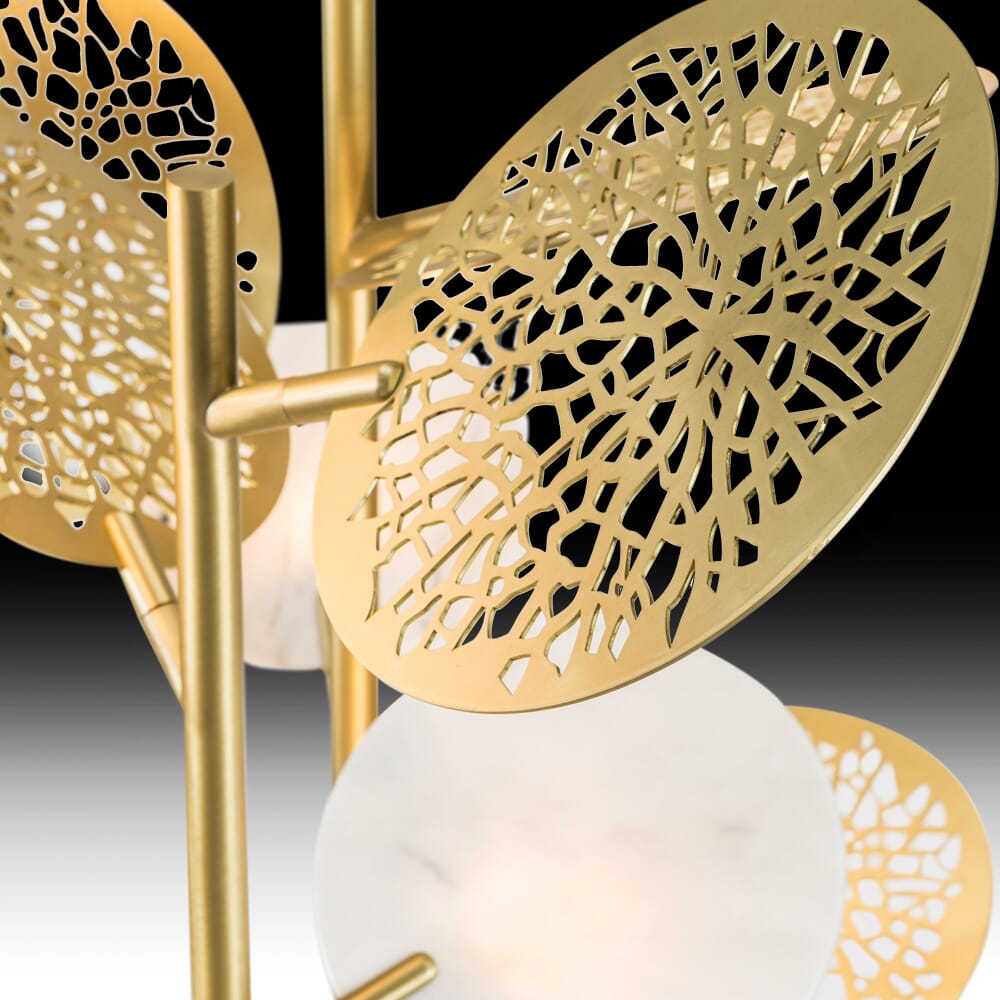 Close-up image of a decorative lamp from a luxury furniture collection featuring golden metal discs with intricate cut-out patterns resembling tree branches. The discs are mounted on a sleek, modern metallic frame with white frosted glass elements in the background. The backdrop is gradient black.