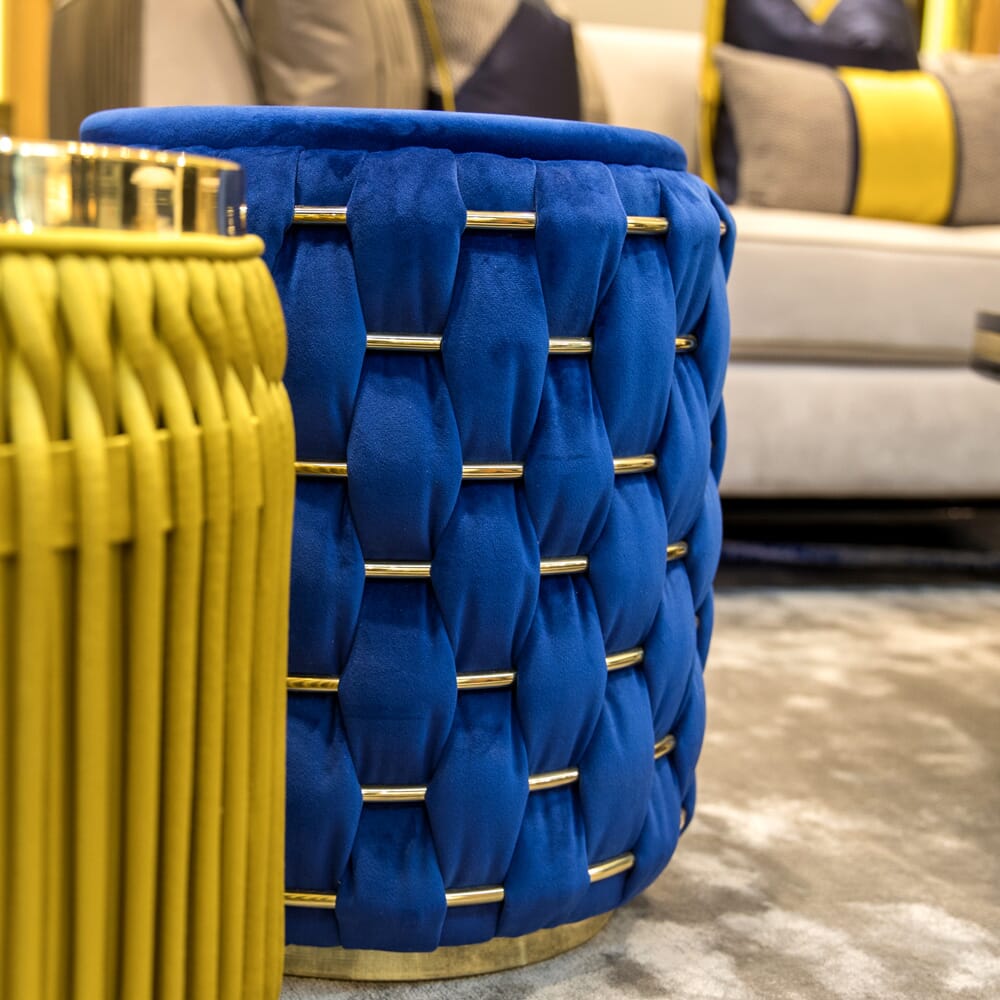 A vibrant blue tufted ottoman with gold accents stands on a gray carpet, part of a luxury furniture collection. Next to it, a yellow woven table is partially visible. In the background, a beige couch with decorative pillows in shades of blue, yellow, and beige creates a harmonious setting.