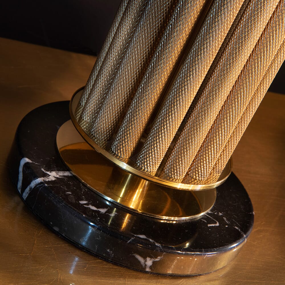 A close-up of the base of a lamp with a textured, cylindrical, golden stand. The base is a combination of gold and black marble, featuring white veins. The lighting highlights the intricate details and rich textures of the materials, making it a standout piece in any luxury furniture collection.