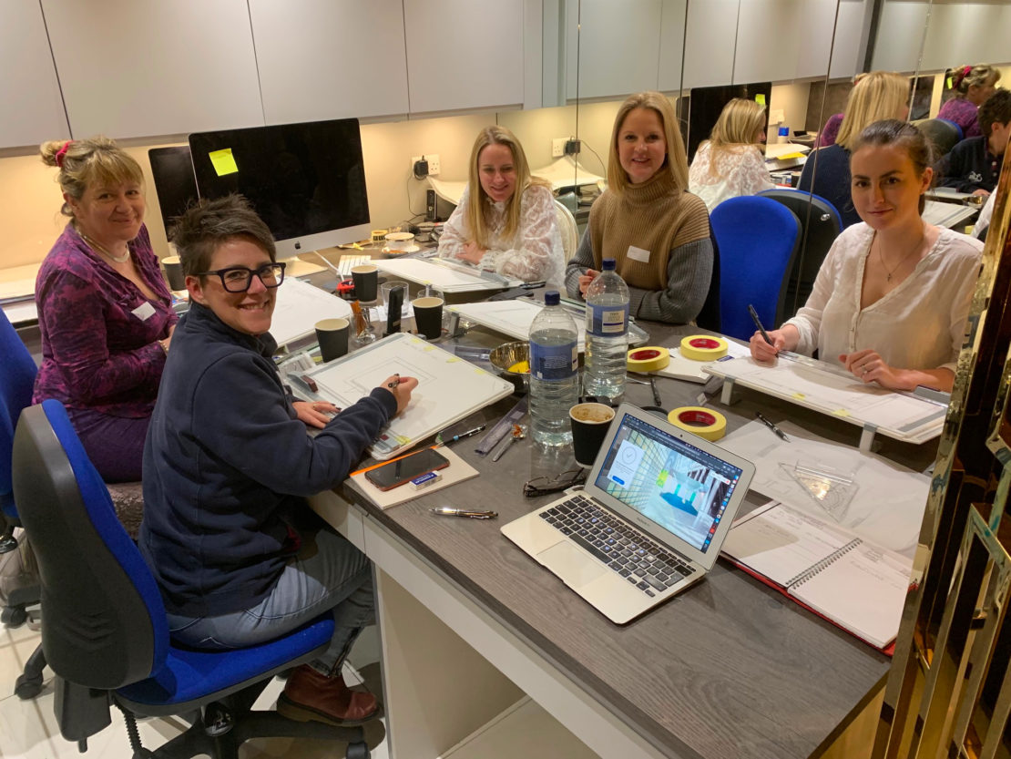 Five women are seated around a table in an office, engaged in work and smiling at the camera. The table has art supplies, a laptop, and several notebooks. The room features a modern design with computer workstations and shelves in the background, reminiscent of an Interior Design Course 2019 setup.