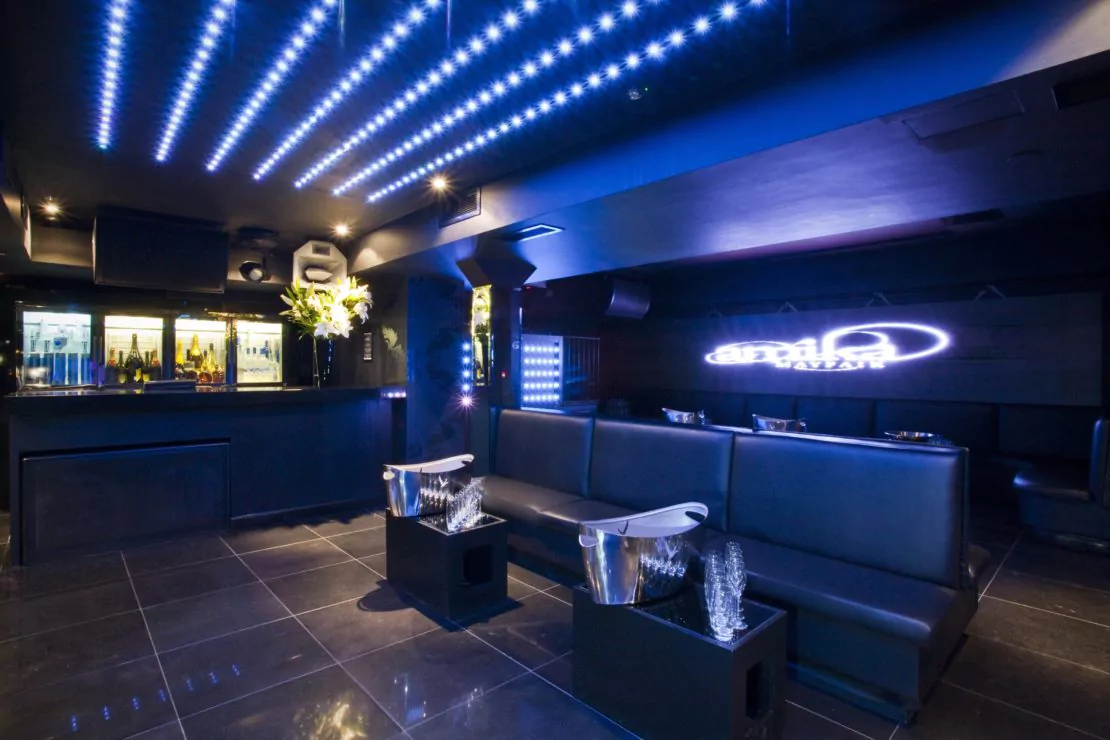 A sleek, modern nightclub interior with black leather seating, illuminated ceiling lights, and a backlit bar area. Tables with ice buckets and glasses are set up, and a neon sign glows on the wall. The space has a sophisticated and stylish ambiance.