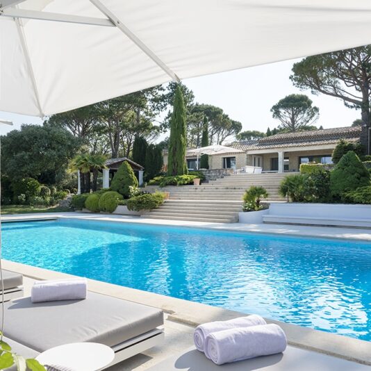 A luxurious outdoor pool with clear blue water, surrounded by cushioned lounge chairs under large white parasols. The pool area is landscaped with lush greenery and leads up to a large, elegant house with multiple levels and tall trees in the background.