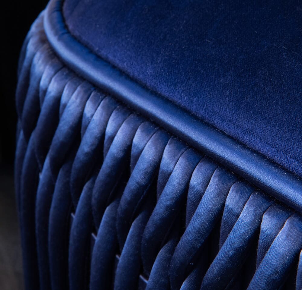 Close-up of a dark blue, velvet-upholstered seat with intricately woven detailing along the side. The fabric has a rich and smooth texture, and the braided design adds a touch of elegance to this piece from our luxury furniture collection.