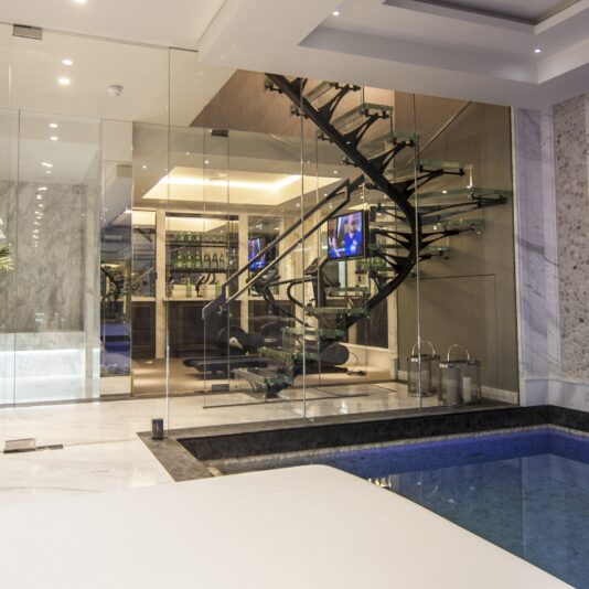 Luxurious modern indoor space featuring a glass-enclosed staircase with black steps, a small indoor pool with a white platform beside it, and sleek marble walls. Tropical plants add greenery, and a large, dark privacy screen covers a window.