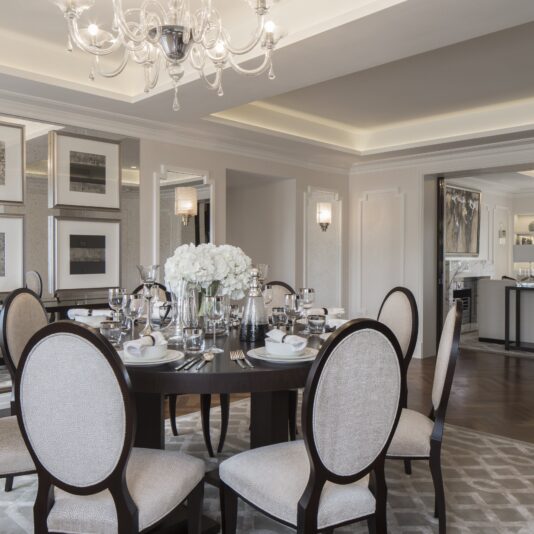 A formal dining room with a round wooden table set for six, reminiscent of Eaton Square elegance. The table is adorned with white flowers in a vase and surrounded by upholstered chairs. The room features refined decor, a chandelier, mirrored walls, and an adjacent open kitchen with contemporary furnishings.