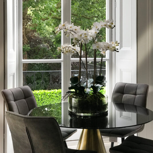 A modern dining area, embodying Knightsbridge interior design, features a round black table with gold accents and four dark grey cushioned chairs. A large pot with white orchids serves as the centerpiece. The space is illuminated by natural light from a floor-to-ceiling window, revealing a leafy green garden outside.