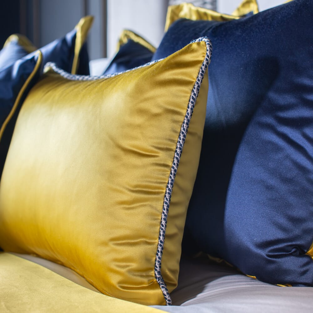 A close-up of a bed with yellow and dark blue satin pillows. The pillows have a shiny finish and contrasting piping along the edges, enhancing their luxurious appearance. Part of our luxury furniture collection, soft lighting highlights the smooth texture of the fabrics.