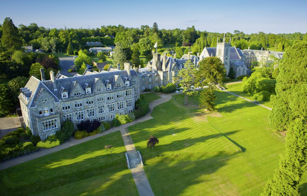 Aerial view of an old stone mansion surrounded by lush green lawns and tall trees. The building features a series of steep, pitched roofs, chimneys, and arched windows. Pathways lead through manicured gardens, offering the perfect setting for a luxury staycation amidst a dense forest backdrop.