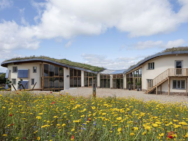 A modern, eco-friendly building with green roofs and solar panels set in a field of blooming wildflowers under a blue sky with scattered clouds. Offering the perfect luxury staycation, the structure has large glass windows and curved roofs, blending harmoniously with the natural surroundings.