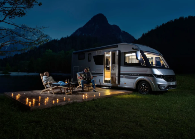 An RV is parked beside a deck in a scenic mountain setting at dusk. Two people are lounging on chairs on the deck, surrounded by lit candles, enjoying the tranquil evening. The RV lights are on, casting a warm glow in the serene outdoor environment of their luxury staycation.