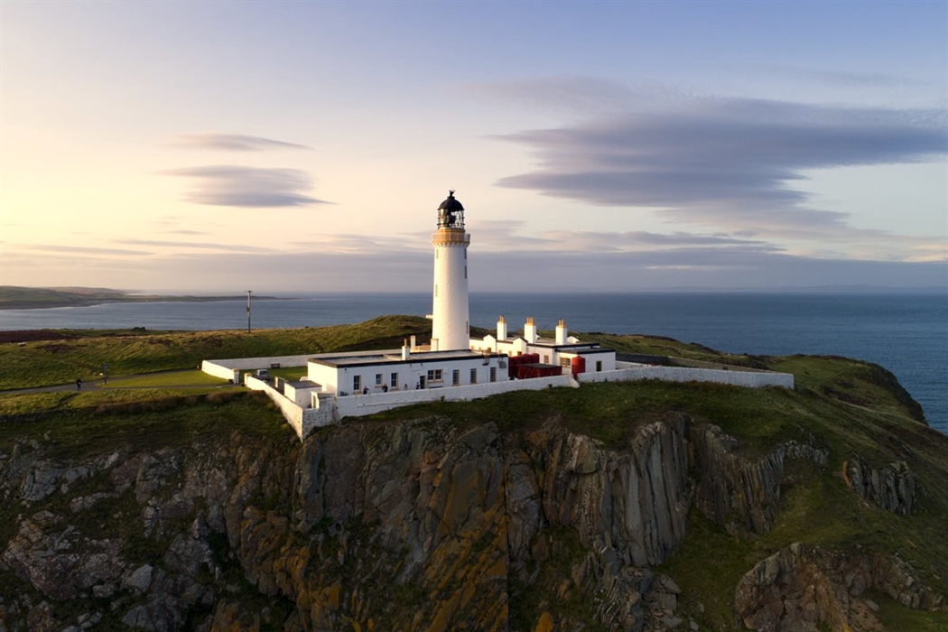 A lighthouse with a white tower and buildings situated on a grassy, rocky cliff by the ocean during sunset offers an exquisite luxury staycation. The sky is clear with scattered clouds, and the surrounding landscape includes distant water and shoreline.