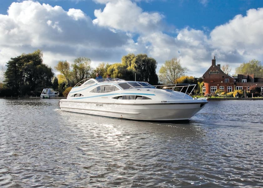 A sleek, white motorboat cruises on a calm river under a partly cloudy sky. Trees with autumn foliage line the riverbank, alongside a picturesque brick building with a tall chimney. Another boat is visible in the distance, hinting at a relaxing luxury staycation.