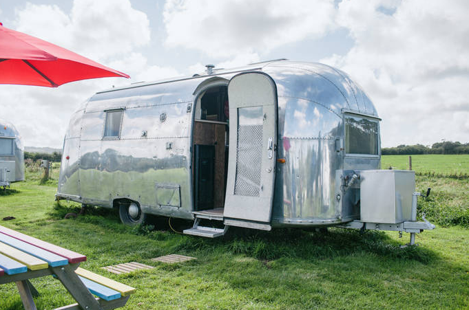 A shiny silver Airstream trailer sits on a grassy field with a partly open door, revealing its luxurious interior, perfect for a staycation. A picnic table with colored slats and a red umbrella stand in the foreground. The sky is partly cloudy.