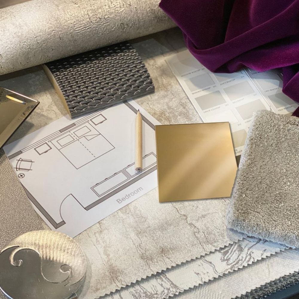 A flat lay image showing interior design materials and tools. There's a floor plan labeled "Bedroom," a pencil, fabric samples in gray and purple, a color swatch, a gold-colored tile, and various textured materials in shades of gray and beige spread on a surface for an interior design course online.