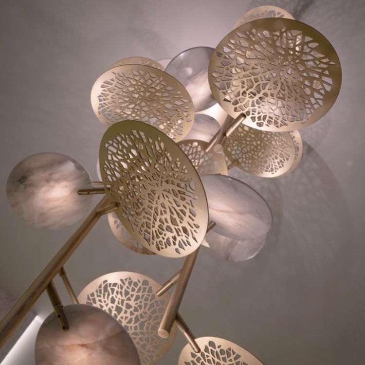 A close-up view of a modern light fixture from the new collection, featuring intricate, circular, metallic designs that resemble lacy leaves and translucent white discs. The light casts delicate shadows on the ceiling, creating an elegant and artistic ambiance.