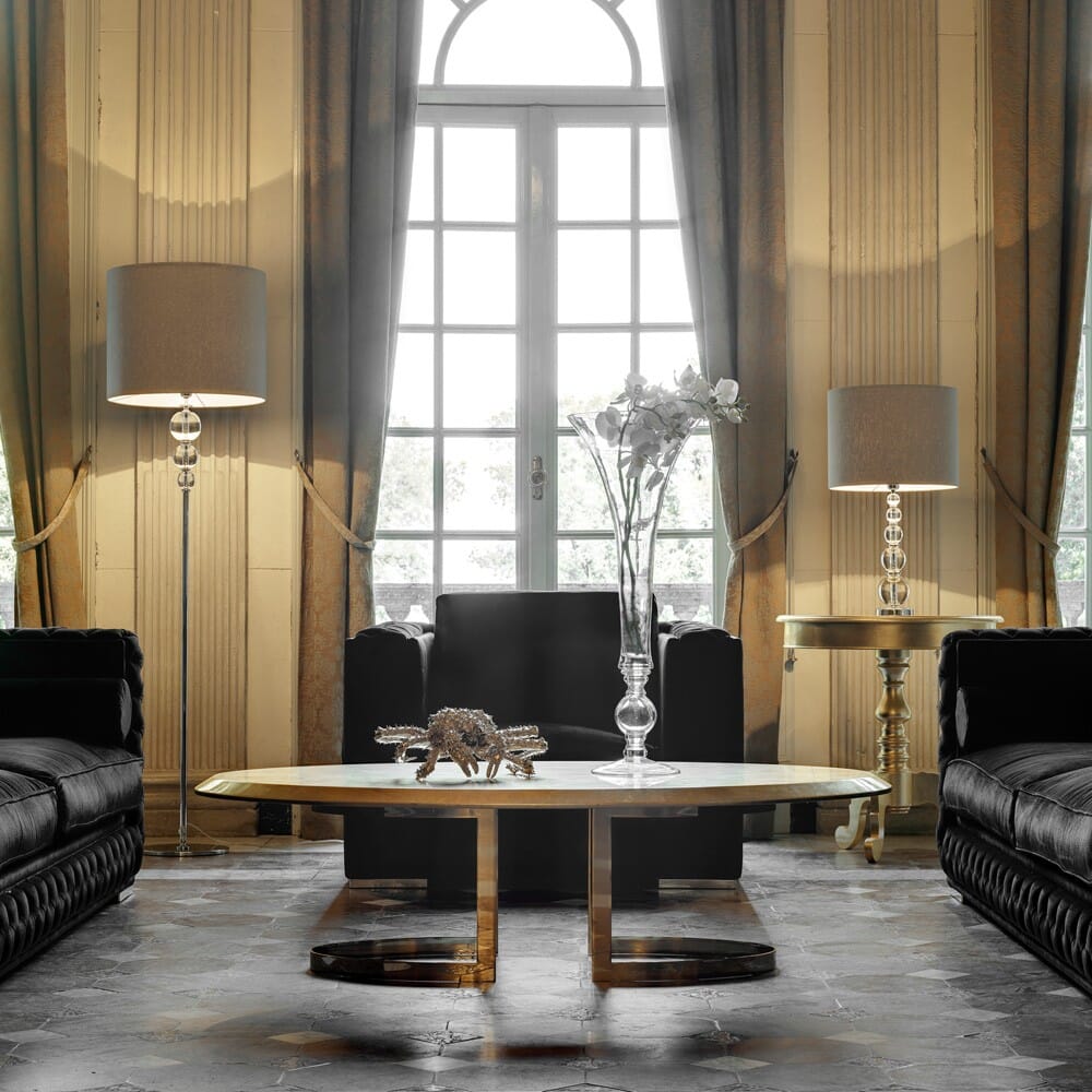 A sophisticated living room reflects the latest interior design trends with two black leather sofas facing each other, a marble and gold coffee table in between. Tall floor lamps stand beside the sofas, while a large window with draped curtains allows natural light to brighten the elegant space.