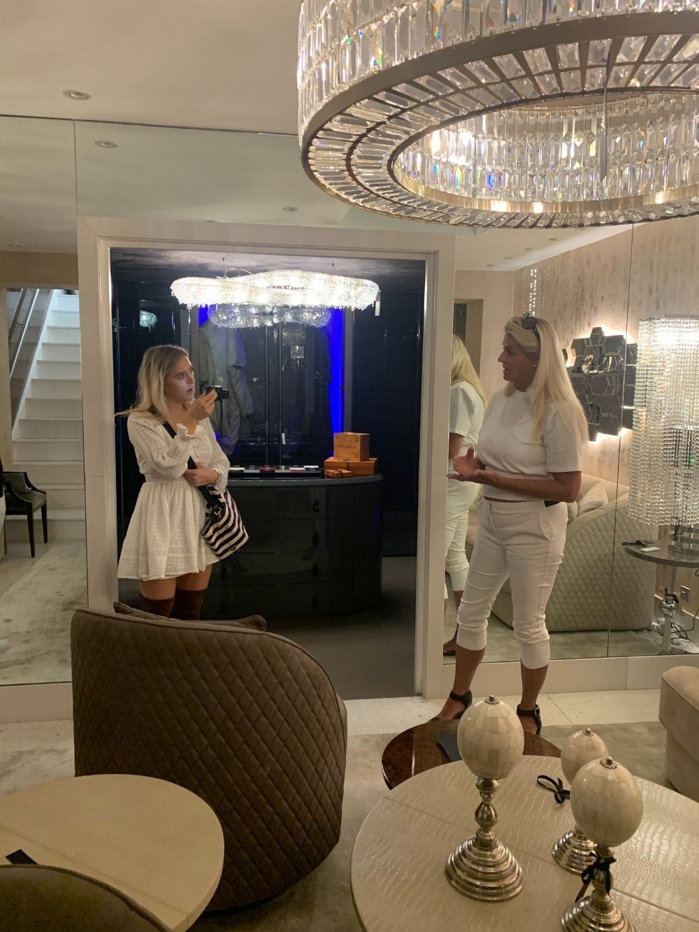 Two women are engaged in conversation in a modern, elegantly decorated living space. The room features a large, ornate chandelier, mirrored walls, and contemporary furnishings from the new collection. One woman wears a white outfit and the other a white dress, holding a striped bag.