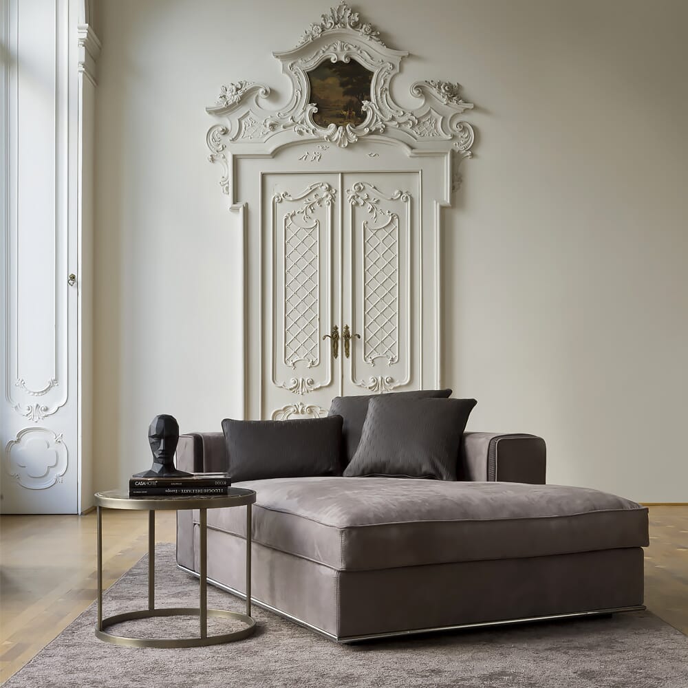 A sophisticated living room, reflecting the latest interior design trends, features an ornate white door with intricate detailing, a large, plush gray chaise lounge topped with gray cushions, a round side table with a black sculpture head and stacked books, and a soft, neutral-toned area rug.