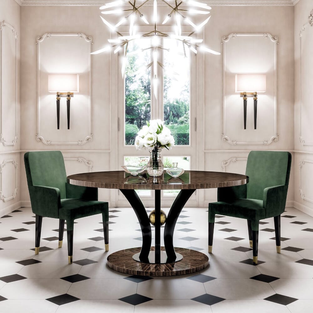 A luxurious dining room featuring a round wooden table with a dark, elegant base. Reflecting the latest interior design trends, two green upholstered chairs sit opposite each other. A bouquet of white flowers adorns the table, under a modern chandelier. Ornate white walls complement the geometric floor tiles.