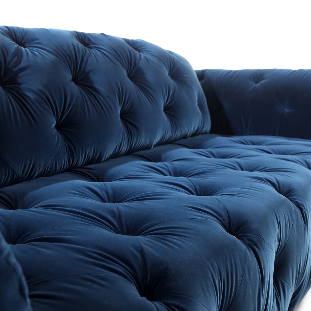 A close-up view of a luxurious, deep blue, tufted velvet sofa showcases the latest in interior design trends. The soft, plush upholstery features a classic button-tufted design that creates a cozy and elegant appearance. The details accentuate the texture and depth of the fabric.