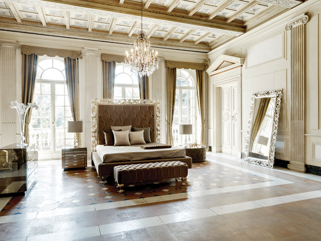 Elegant bedroom featuring a large, tufted bed with luxurious linens, flanked by two modern nightstands with lamps. Embracing interior design trends, the room has tall arched windows with curtains, a grand chandelier, intricate ceiling details, and a large ornate mirror leaning against the wall.