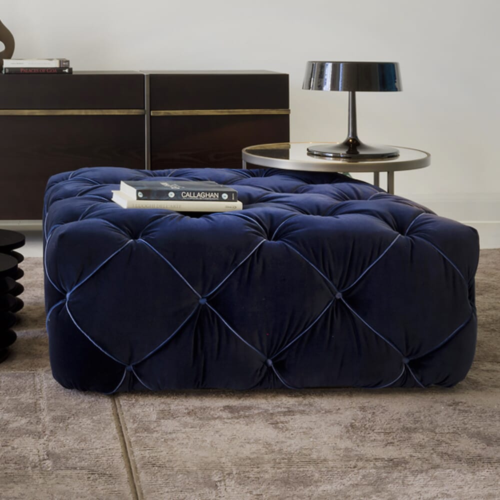 A luxurious dark blue, tufted ottoman is placed on a light-colored rug. Embracing the latest interior design trends, a round side table sits beside it, holding a modern black lamp and a book titled "CALLAGHAN." In the background, there's a dark wood console against a white wall.