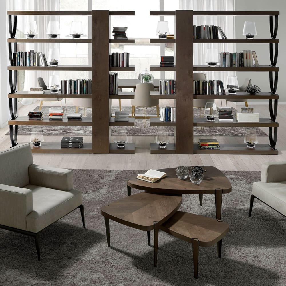 Modern living room with neutral tones featuring two comfortable armchairs, a unique tiered coffee table with a wooden finish, and two large, sleek bookcases filled with books and decorative items. Following the latest interior design trends, the space has a plush gray carpet and minimalistic decor.