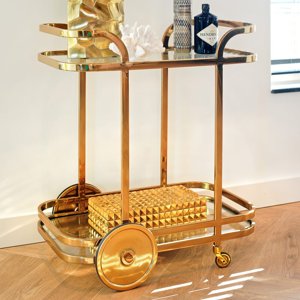 A gold bar cart with two glass shelves stands on a light wooden floor, embodying the latest interior design trends. The cart is adorned with decorative items including a vase, a coral sculpture, and a bottle of Hendrick's Gin. A textured gold object is placed on the lower shelf while sunlight filters through nearby blinds.