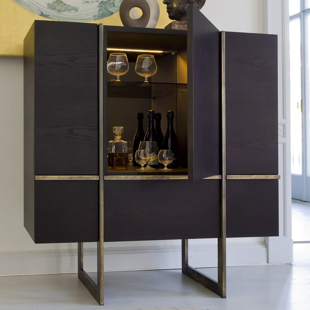 A modern dark wooden cabinet with brass accents stands in a light-colored room, mirroring the latest interior design trends. Its doors are partially open, revealing shelves with wine glasses, decanters, and bottles inside, illuminated by subtle interior lighting. The base of the cabinet is metallic.