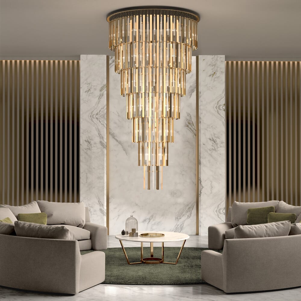 A luxurious living room that follows the latest interior design trends features a grand, multi-tiered chandelier with cylindrical lights hanging above a white marble coffee table. The table is set on a dark green rug, surrounded by four beige armchairs. The background boasts vertical wood and marble panels.