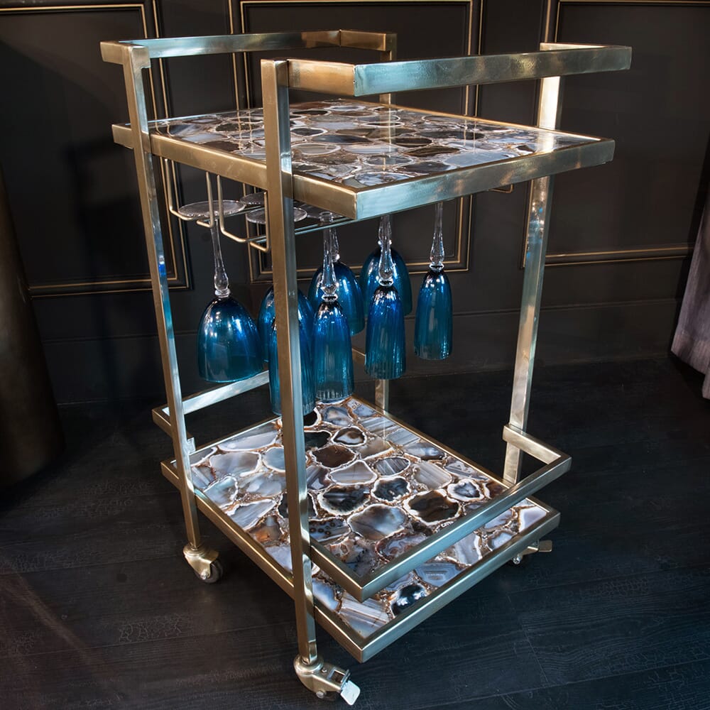 A stylish bar cart with a gold frame and two glass shelves featuring a black and white marbled pattern. Blue wine glasses hang upside down from the upper shelf. The cart, aligning with current interior design trends, has wheels for easy movement. The background includes dark walls with minimal detailing.