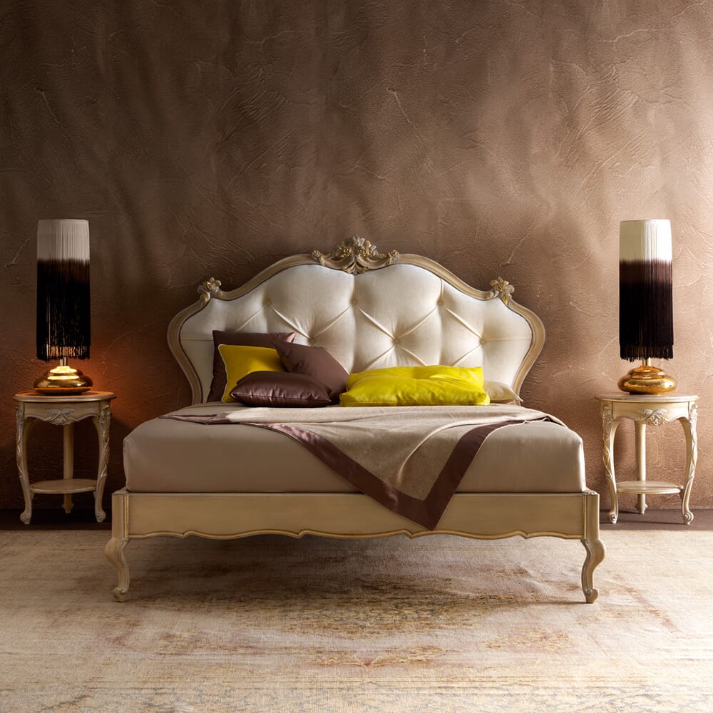 Luxurious bedroom with an ornate beige and white upholstered bed frame set against a textured brown wall. Two wooden nightstands, each with a decorative lamp featuring a gradient black and white shade, flank the bed. Embracing interior design trends, the bed is made with brown, yellow, and white bedding.