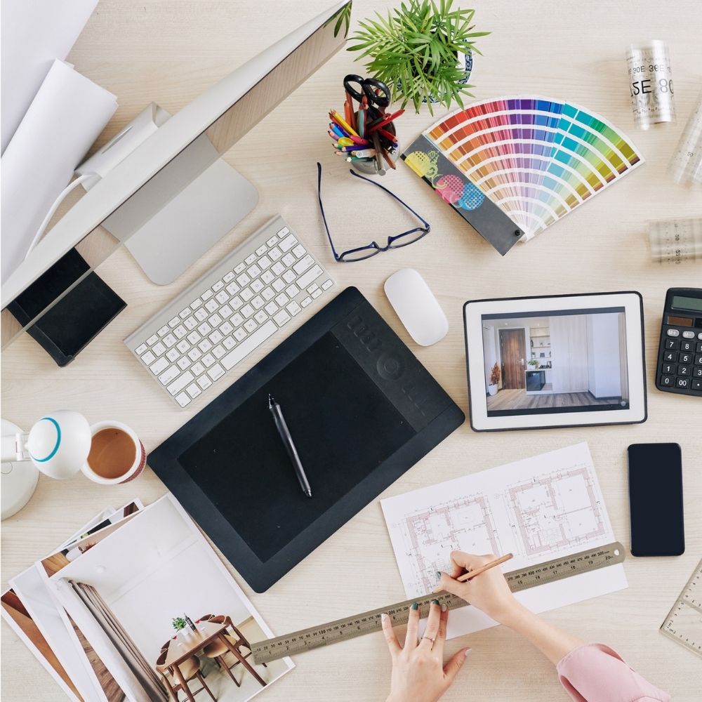 A cluttered desktop features an architectural workspace with a keyboard, digital drawing tablet, computer monitor, color swatches, miniature model, blueprints, ruler, coffee cup, plant, notepad, glasses, various stationery items—and materials for an online interior design course.