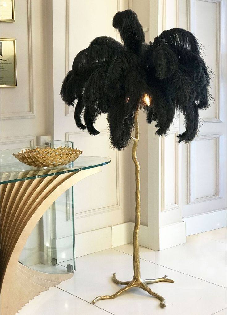 A unique standing lamp with a gold-colored, branch-like base and a shade made of black feathers makes for the perfect luxury gift. The lamp is placed next to a glass table with a decorative gold bowl on it, within a stylish, white-walled interior setting.