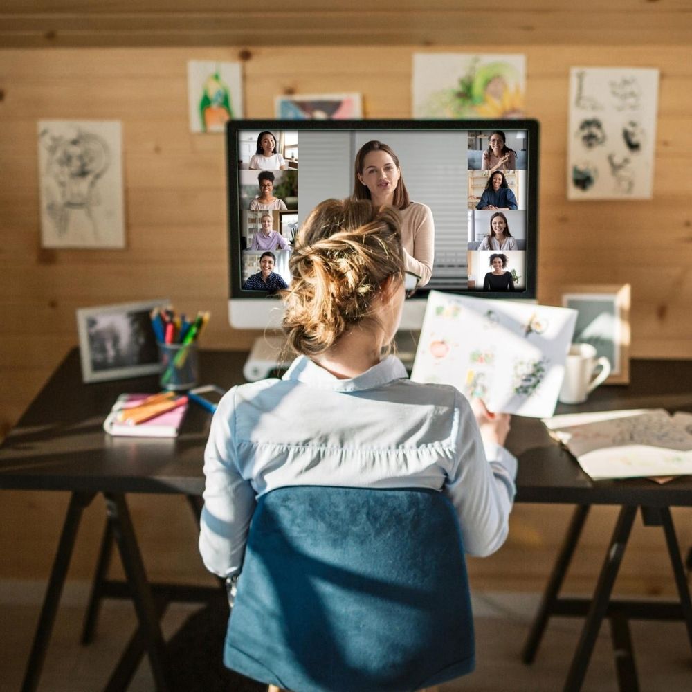 A person with light brown hair in a bun sits at a desk with art supplies, looking at a computer screen displaying a video conference with five people. The wooden wall behind the desk is adorned with various sketches and drawings, creating an atmosphere as cozy and inspiring as unwrapping a luxury gift.