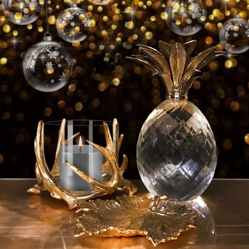 A festive still life features a faceted crystal pineapple with gold leaves, a lit candle in a glass holder with a gold antler base, and decorative gold leaves. Snowflake-adorned glass ornaments and golden bokeh lights create a warm, celebratory Christmas decor backdrop.