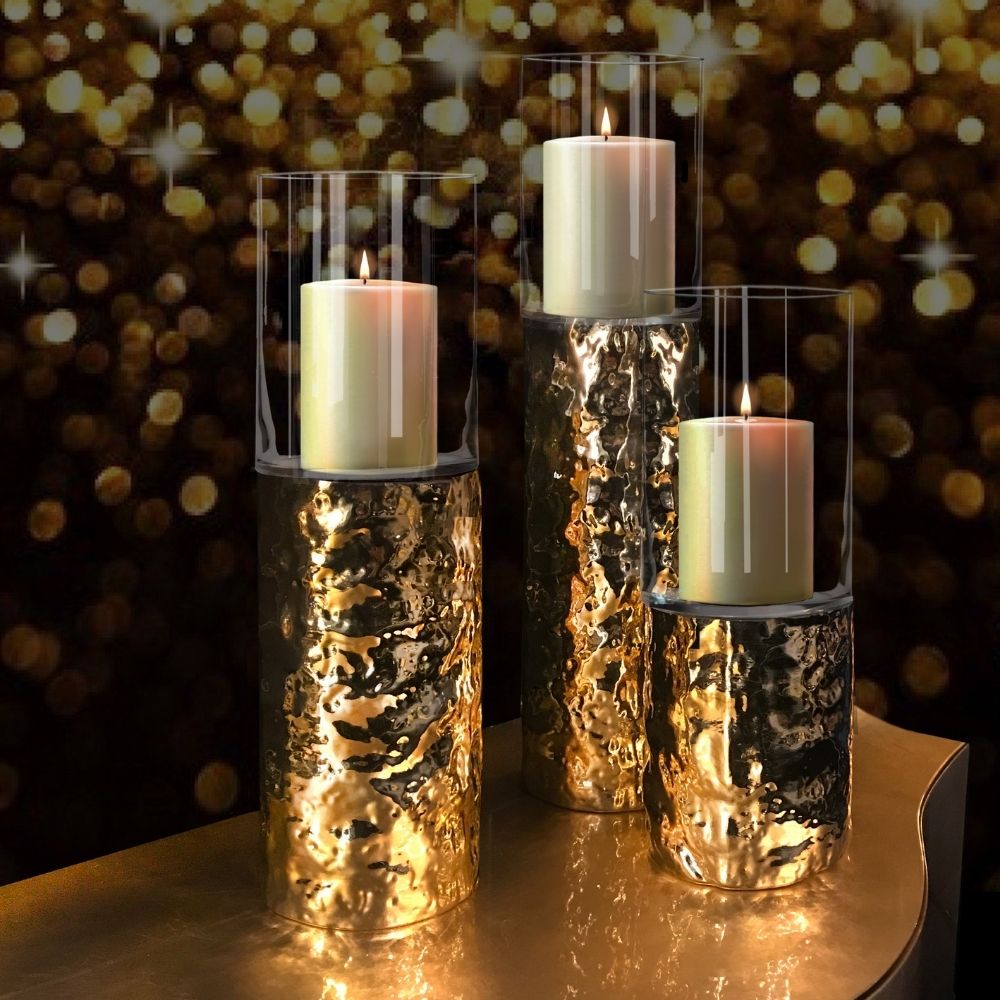 Three pillar candles in textured, reflective golden holders topped with clear cylindrical glass coverings are lit, casting a warm glow. The background features a bokeh effect of sparkling golden lights, creating a festive and elegant Christmas decor atmosphere.