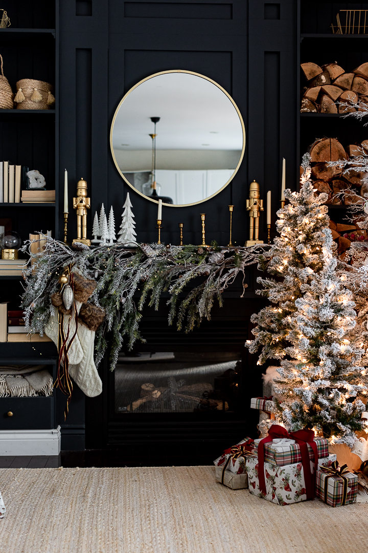 A cozy living room decorated with Christmas decor features a dark fireplace with a round mirror above it. Garland, stockings, and candle holders adorn the mantel. A lit, snow-covered Christmas tree stands nearby with wrapped gifts underneath. Shelves hold books and logs.