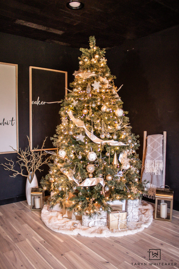 A beautifully decorated Christmas tree stands in a corner of a dark-themed room. The tree is adorned with gold and white ornaments, ribbons, and twinkling lights. Wrapped gifts rest beneath on a plush white tree skirt, enhancing the Christmas decor. A blanket drapes over a chair nearby.