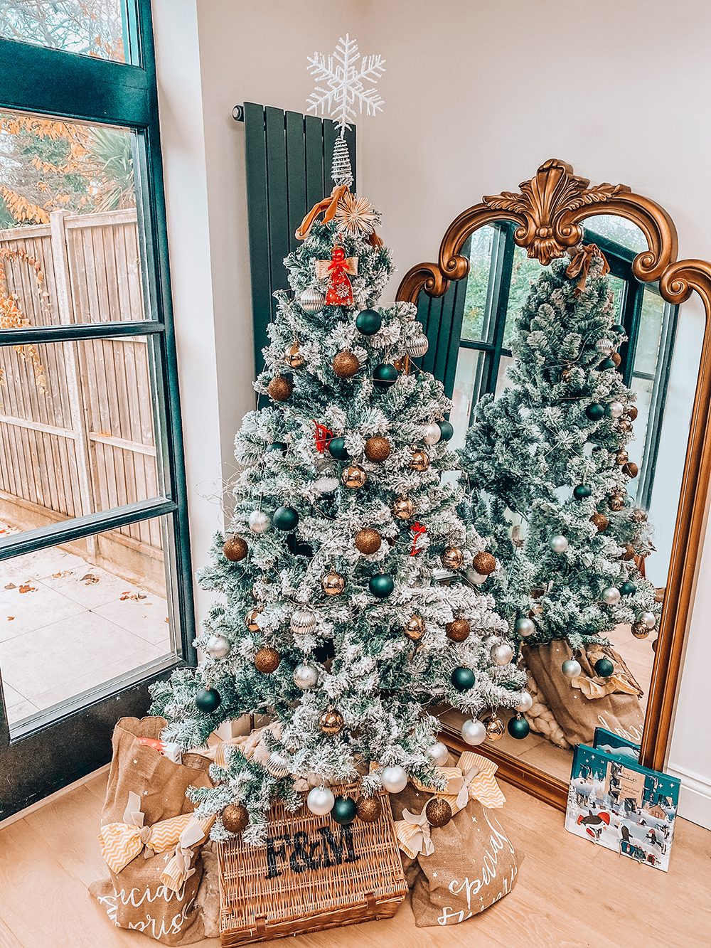 A decorated Christmas tree with a mix of green, brown, and white ornaments stands in front of a large ornate mirror. The tree is topped with a white snowflake. Reflecting in the mirror, this charming piece of Christmas decor is surrounded by wrapped presents and has a basket underneath labeled "TIDAL.