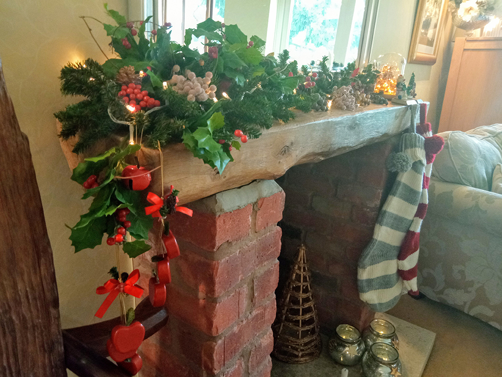 A cozy brick fireplace is adorned with Christmas decor, featuring a garland of evergreen branches, holly, berries, and pinecones. Red ribbon ornaments and a hanging striped stocking add festive cheer. Fairy lights and decorative jars brighten the scene.