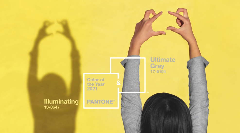 A person forming a heart shape with their hands in front of a yellow background casts a shadow. PANTONE’s 2021 Colors of the Year, Illuminating 13-0647 (yellow) and Ultimate Gray 17-5104, are featured with labels positioned over their respective colors in the image.