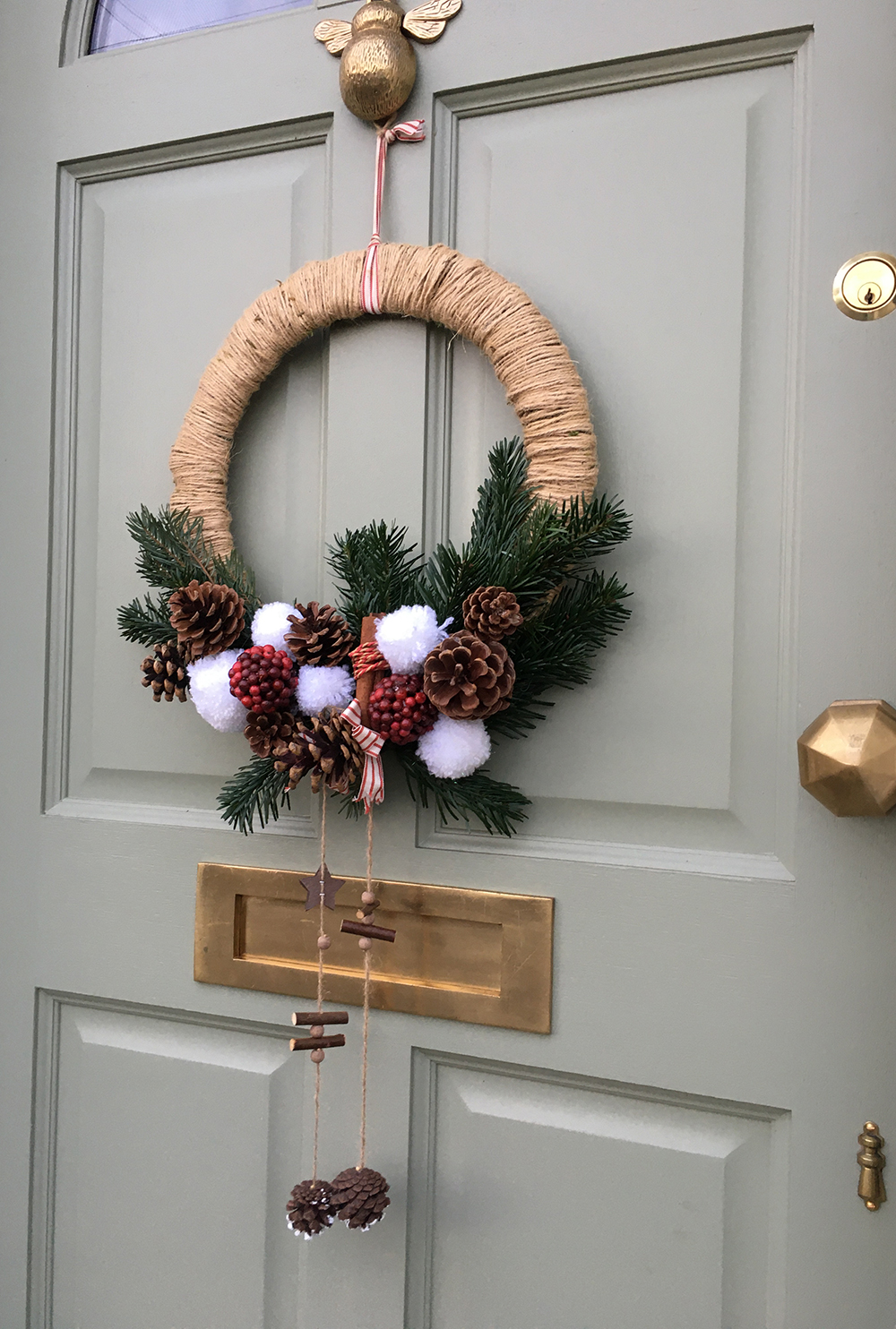 A green door adorned with rustic Christmas decor: a holiday wreath made of wrapped twine, decorated with pinecones, white faux snowballs, and red berries. Hanging from the wreath are wooden stars and pinecones. The door has a brass knocker and mail slot.