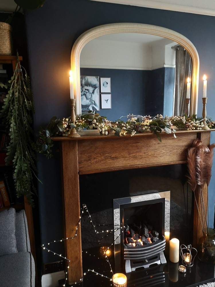 A cozy living room scene with a dark blue accent wall and a wooden fireplace mantel adorned with Christmas decor, greenery, and white lights. A large mirror with candles on the mantel reflects part of the room. The fireplace is lit, and the space feels warm and inviting.