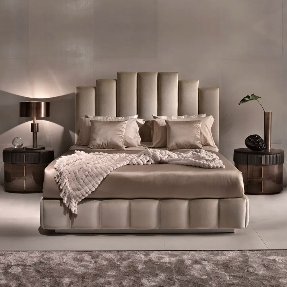 A modern luxury bedroom interior design features a large upholstered bed with plush beige pillows and a matching bedspread. Two round bedside tables with lamps flank the bed. A small plant decoration and a sculptural art piece adorn the tables, blending seamlessly with the monochromatic decor.