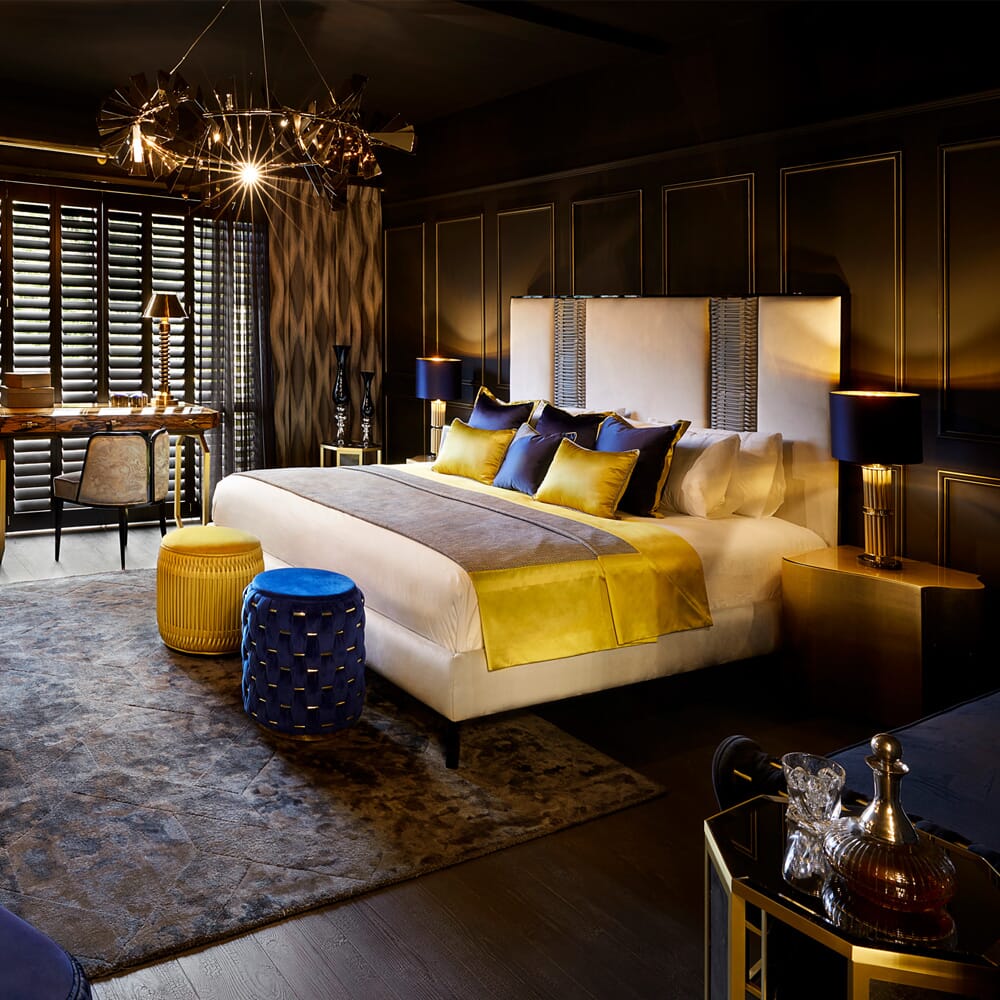 A luxury bedroom interior design featuring a large bed with yellow and blue pillows and a yellow throw. Stylish lamps adorn bedside tables, while an elegant chandelier hangs overhead. There's a desk by the window, patterned walls, a plush rug, and gold and blue accents throughout the room.