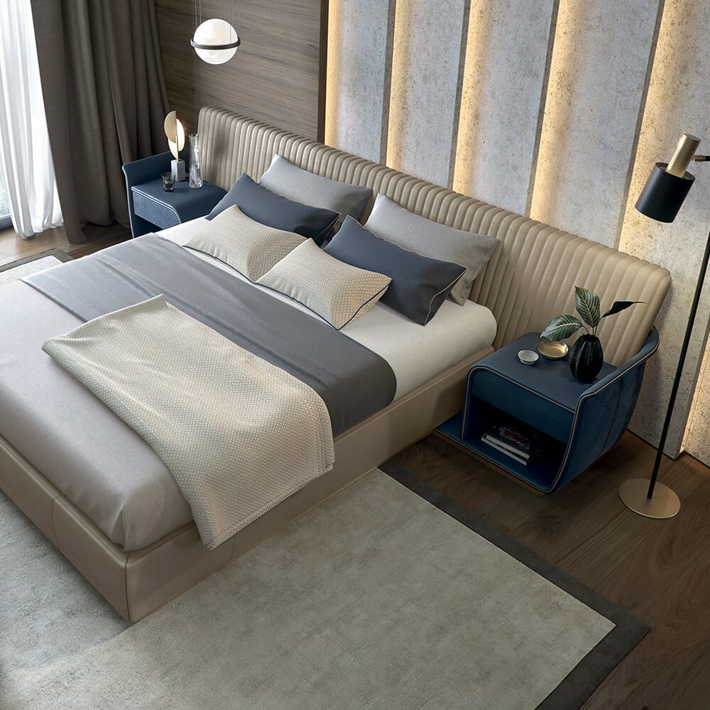 A luxury bedroom interior design features a beige upholstered bed with gray and beige bedding. Two blue nightstands with potted plants, lamps, and decorations flank the bed. The room is accented with a textured wall, floor lamp, and a large window with curtains on the left.