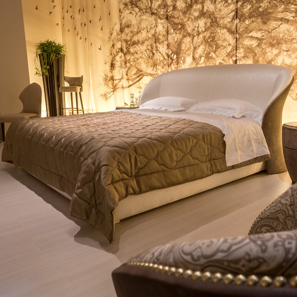 A warmly lit luxury bedroom with a queen-sized bed featuring a light brown quilted bedspread and white pillows. Behind the bed is a decorative wall with a tree design. The room boasts elegant beige and brown furniture, including a chair, side table, and plant, showcasing impeccable interior design.