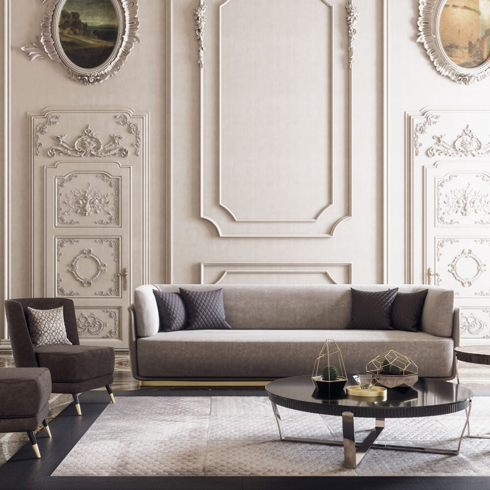 A luxurious living room features an ornate wall with intricate mouldings and framed artworks. A taupe sofa with navy throw pillows is centered, flanked by two brown chairs in warm neutrals. A black round coffee table holds decorative geometric sculptures, and a patterned rug covers the floor.