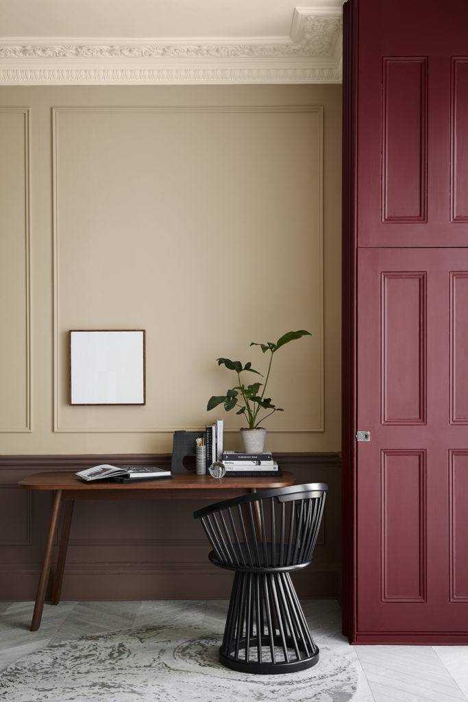 A modern workspace featuring a wooden desk with books, a potted plant, and a clock on it. A black spindle chair is placed in front of the desk. The room showcases warm neutrals with its beige paneled walls, white ceiling with intricate molding, and a maroon door to the right.